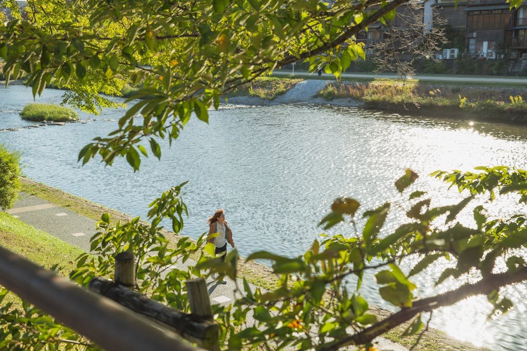 Anonymous woman walking on embankment near river in green park in sunlight