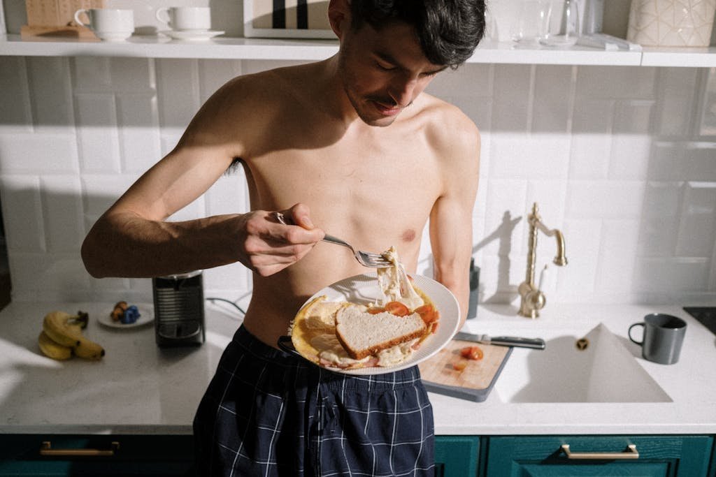 Topless Man in Blue and White Plaid Shorts Holding Stainless Steel Fork and Knife Slicing Cake