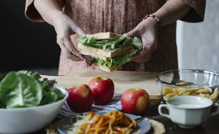 Unrecognizable female standing at table with apples and cutting board with sandwich with lettuce in hands in kitchen on breakfast time