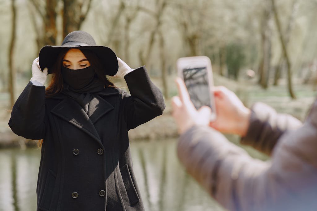 Couple in warm coat wearing latex gloves and medical mask taking photo in park near pond and trees during coronavirus epidemic