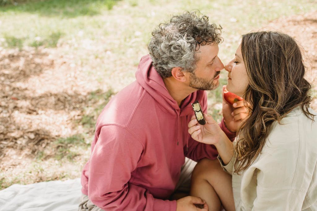 Side view of enamored adult couple in casual clothes eating delicious snacks and preparing to kiss during picnic in nature on sunny day