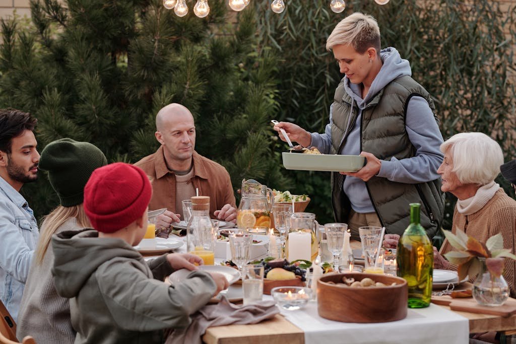 Smiling woman wearing warm vest and hoodie serving baked dish during holiday party at big table under garlands and evergreen trees beside