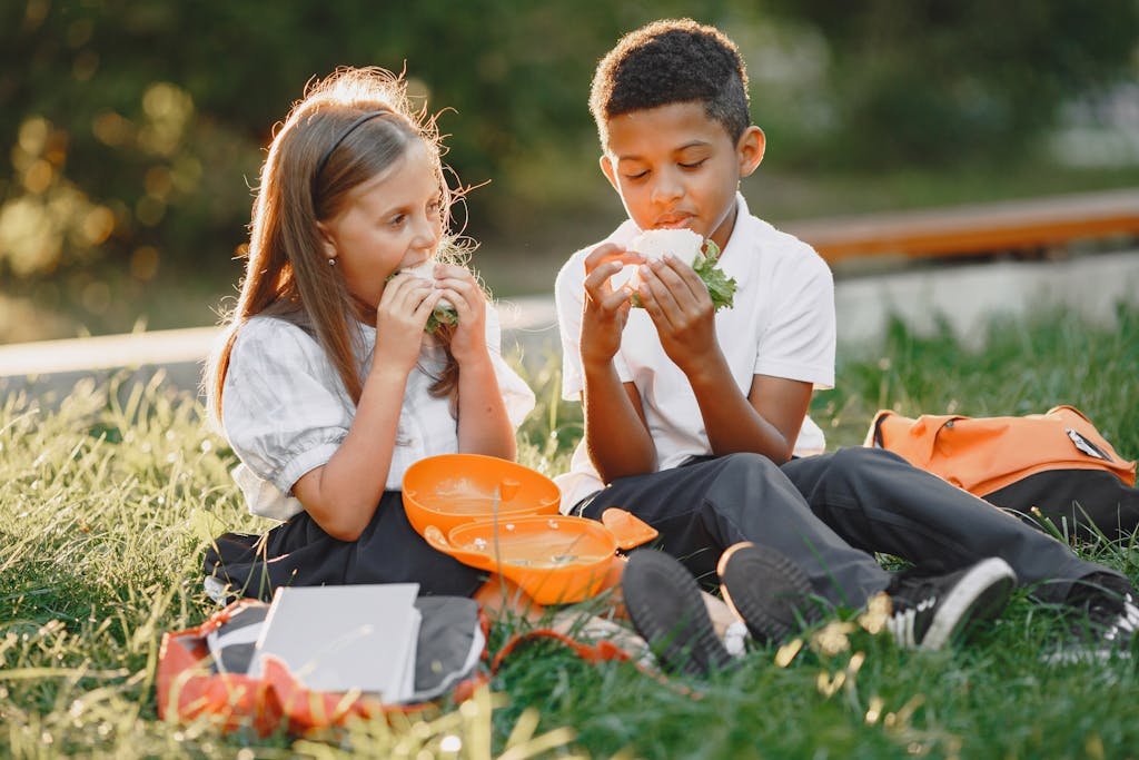 Young Students Eating Sandwiches while Sitting on Green Grass