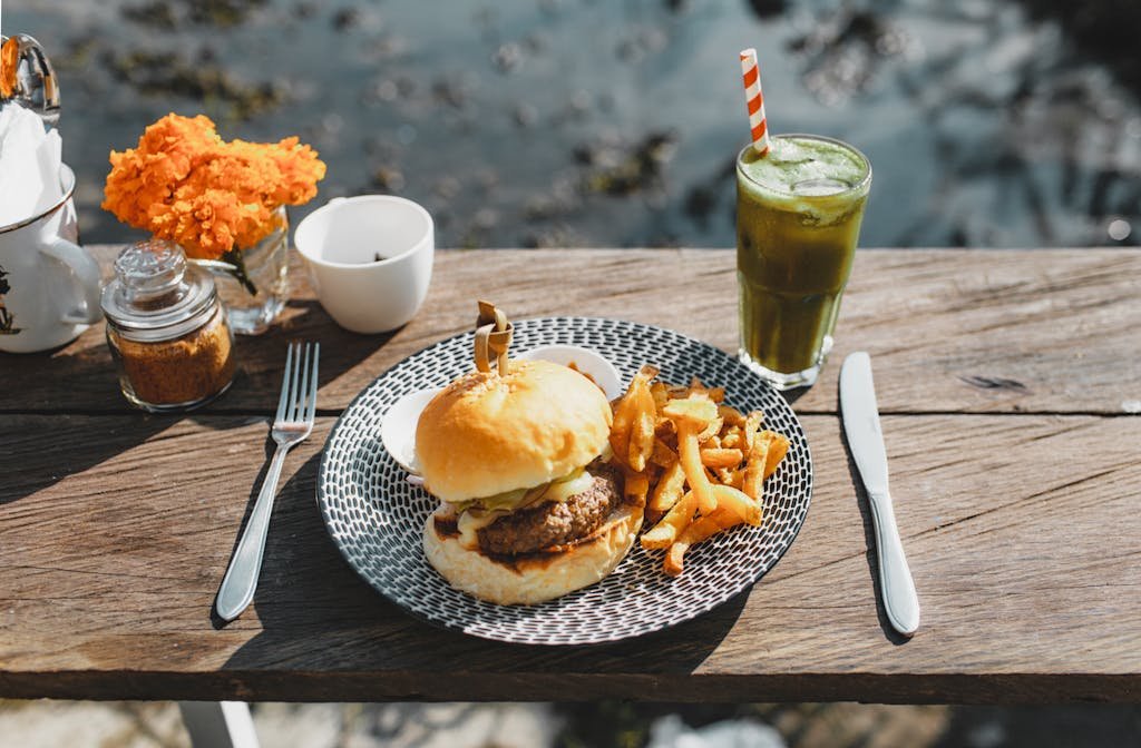 Plate with appetizing hamburger and french fries placed on lumber table near glass of green drink in outdoor cafe