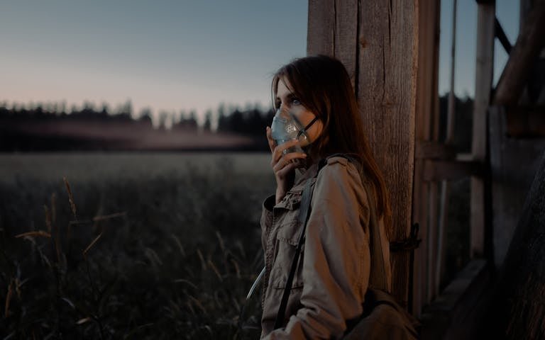 A woman in a brown jacket leans against a wooden column outdoors, wearing a facemask during twilight.