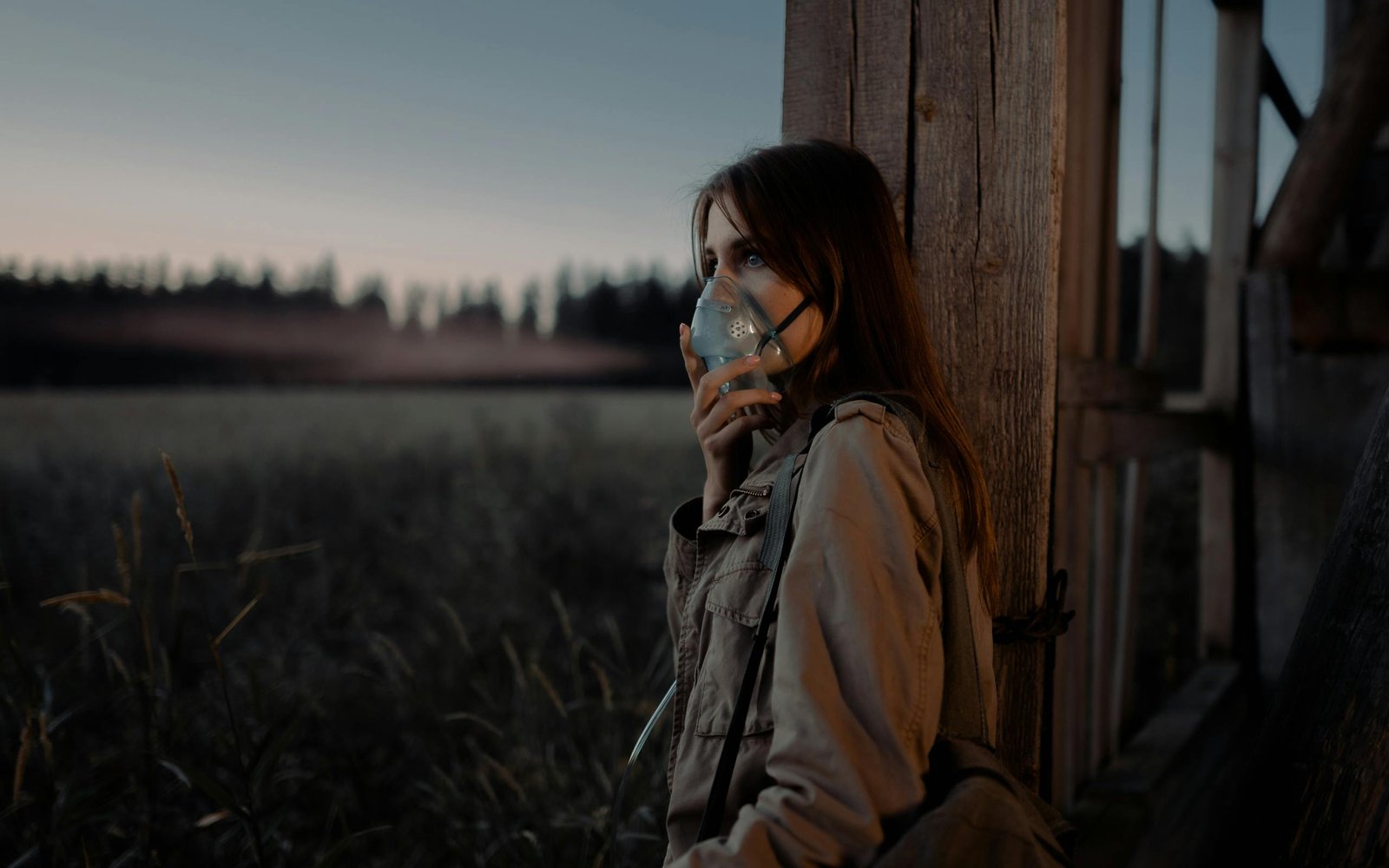 A woman in a brown jacket leans against a wooden column outdoors, wearing a facemask during twilight.