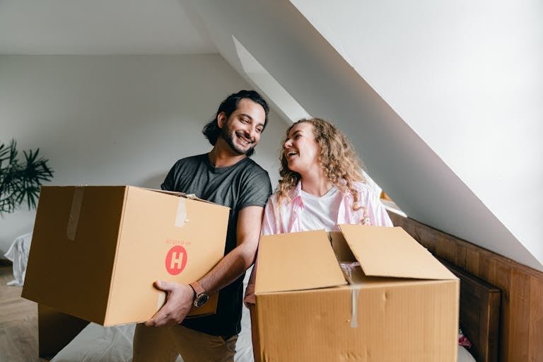 Content young woman and ethnic bearded man carrying cardboard boxes and laughing each other while unpacking belongings in new modern house